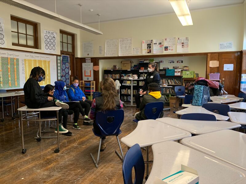 The image shows a classroom setting with students and a teacher. Some students are seated at desks, while others are gathered in a group. The teacher appears to be addressing the students. There are books and other learning materials visible in the background.